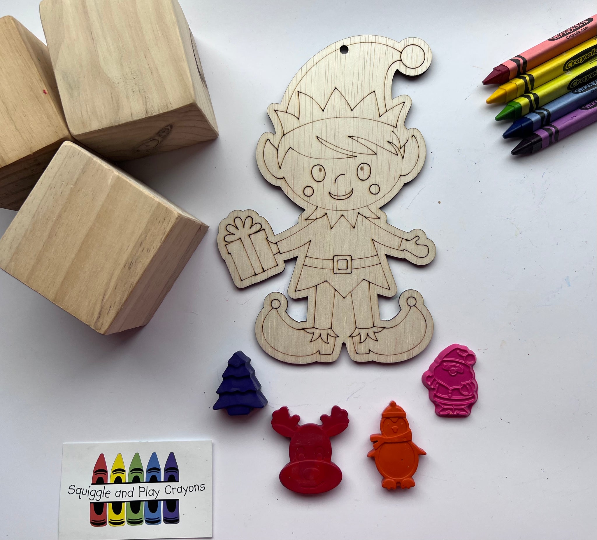 wooden cut out shaped in a male Christmas elf holding a square box present. 4 Small crayons in the shape of a Christmas tree, head of a reindeer, standing penguin wearing a beanie and scarf, standing Santa