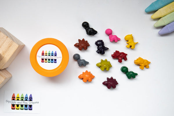 Four small brachiosaurs shaped dinosaur crayons. Two small t-rex shaped dinosaur crayons. Four small stegosaurs shaped crayons. Two small triceratops shaped crayons. Small tub container with Squiggle and Play Crayons logo sticker on the circle shaped lid